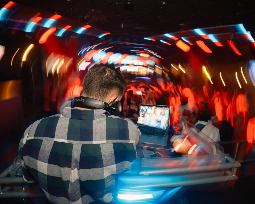 DJ seen from behind, mixing for a nightclub crowd beneath sweeping light trails